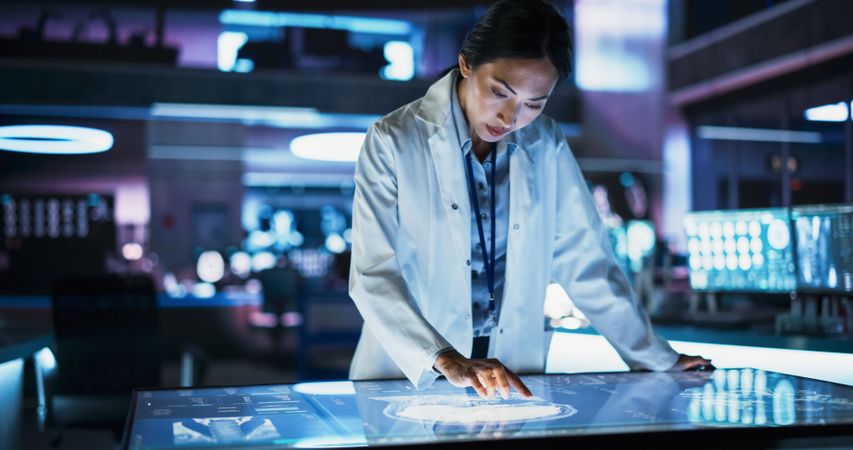 Female Asian Neuroscinetist Using Interactive Touch Screen Table With MRI Scans On Display In Modern Biotechnology Research Center.