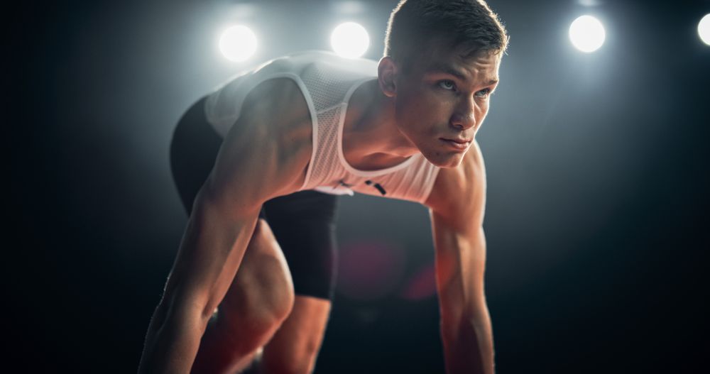 Strong Young Man Starting a Race From Track Starting Blocks Position on a Dark Stadium in the Evening. Portrait of a Fit Male Sprint Runner Participating for an International Competition