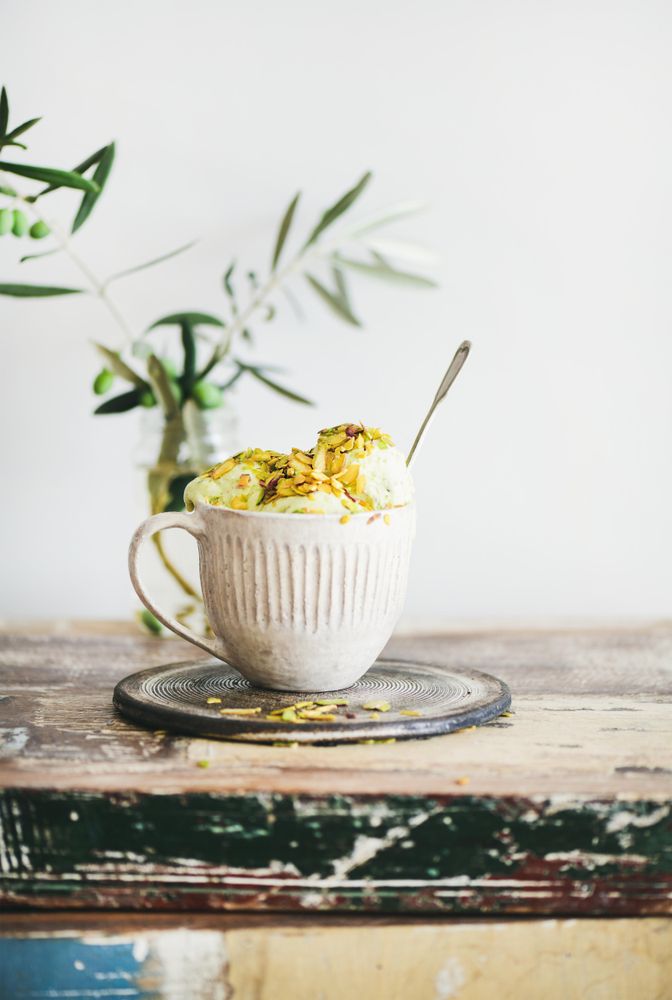 Cup of pistachio ice cream with spoon sticking upright with leaves on light background