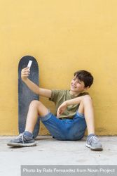 Boy sitting on ground leaning on a yellow wall and taking a selfie, vertical 56Glzj