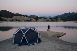 Man with binoculars and dog looking over lake with tent at sunset