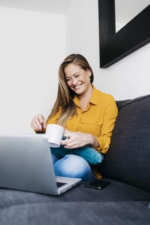 Smiling long haired female sitting on sofa with laptop computer working remotely from home