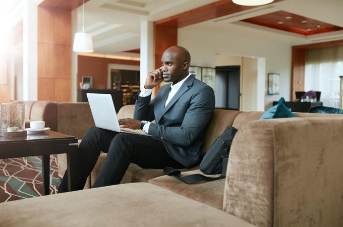 Businessman sitting on sofa with a laptop using mobile phone