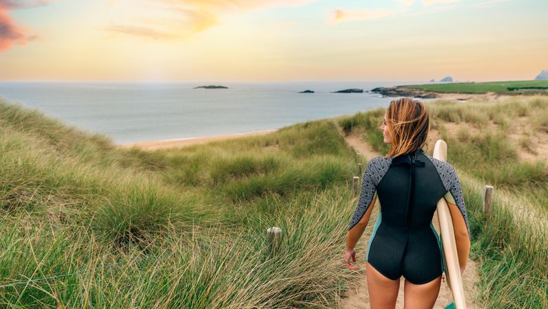 Back of woman standing on cliff overlooking beach holding a surfboard