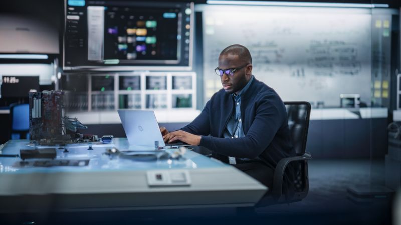 Portrait of a Black Male Specialist Using Laptop Computer to Improve Electronic Circuit Board in Modern Office. African Engineer Thinking and Typing Ideas for Artificial Intelligence Project