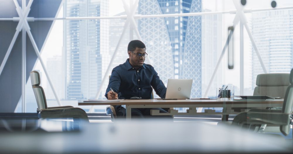 Portrait of Successful Black Businessman Working on Laptop Computer in His Big City Corner Office.