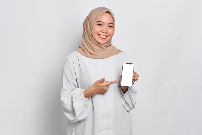 Happy Asian Muslim woman in a bright studio shoot holding cell phone and pointing to screen