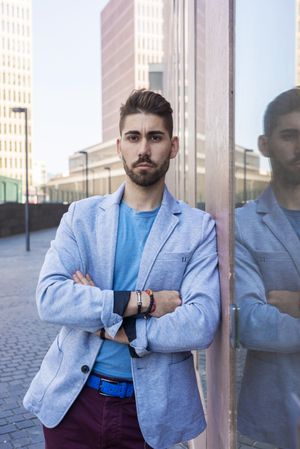 Young man in blazer with arms crossed leaning on a building wall outside looking at camera, vertical