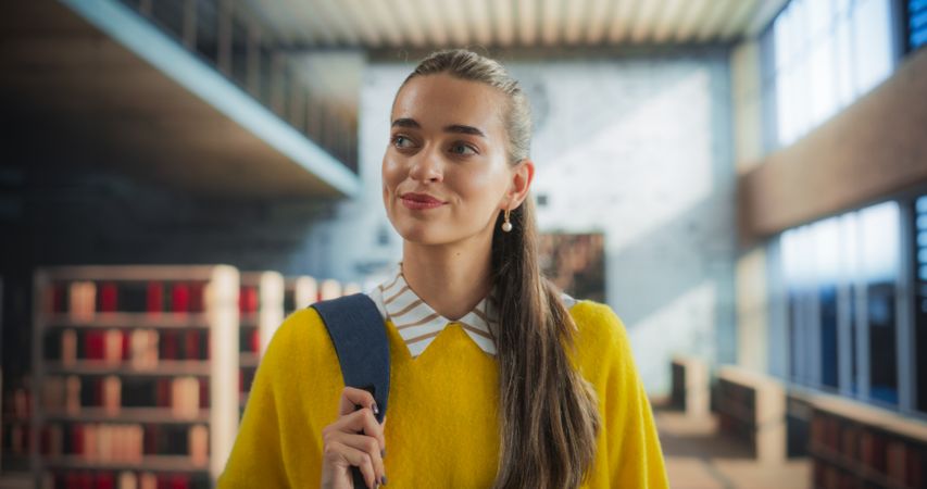 Close Up Portrait of a Happy Smiling Young Student Walking Indoors in University Campus. Joyful Female Wearing a Yellow Jumper, Looking Around the Library, Searching for Textbooks for Lecture