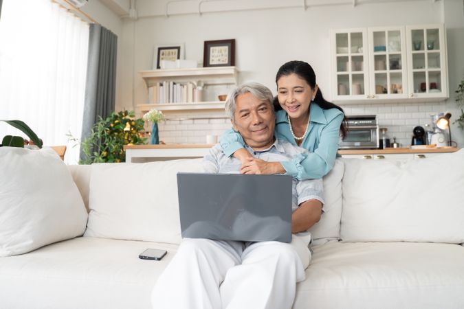Older Asian man sitting on couch working on laptop while his wife embraces him