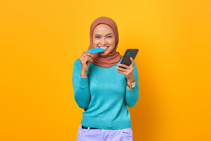 Muslim woman looking happy biting on credit card and holding smartphone