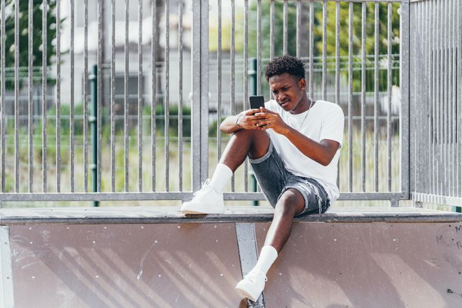 Sportive black men thoughtfully browsing smartphone while sitting on fence