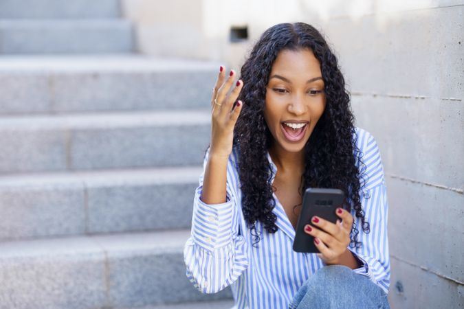 A young and enthusiastic woman is happily engaging with her smartphone while sitting on the steps