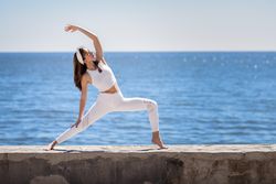 Woman wearing sport clothes and doing warrior A pose with coast in the background