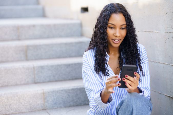 A young woman is engaged with her smartphone while sitting on outdoor steps, smiling happily