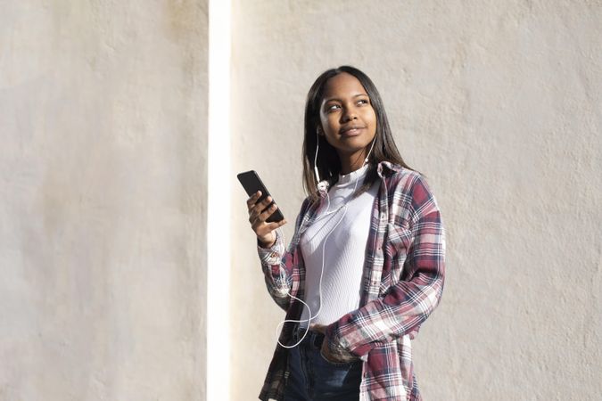 Female standing in the sun in front of wall holding phone and looking around