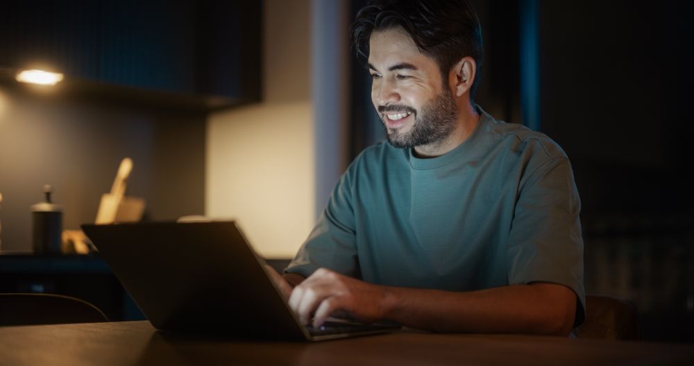 Authentic Latin Man Sitting at a Table in a Cozy Kitchen and Using Laptop Computer at Home at Night.