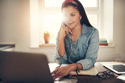 Content woman reading on laptop in home office with earbuds and pencil
