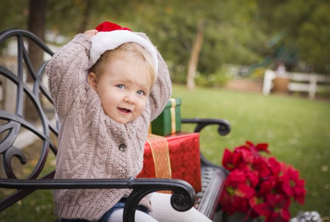Young Child Wearing Santa Hat Sitting with Christmas Gifts Outside.