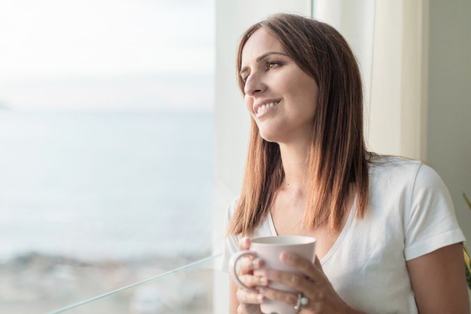 Woman looking out window wistfully while holding cup of coffee