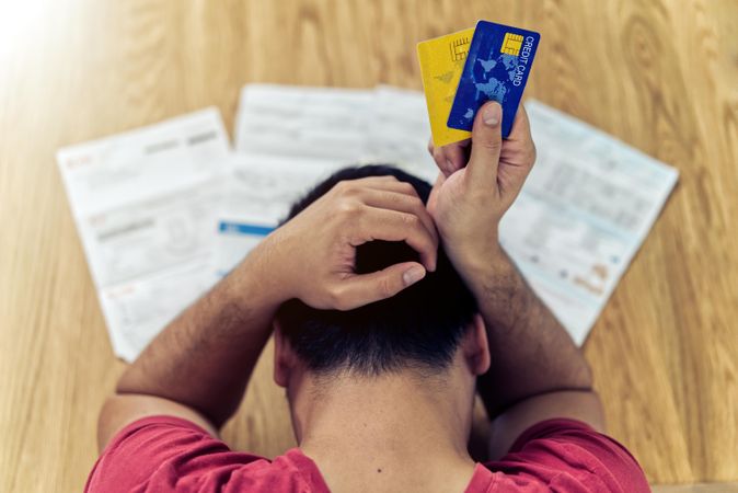 Back view of frustrated man holding credit cards sitting at a table with documents