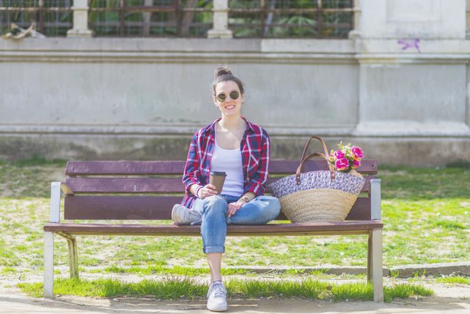 Front view of a young hipster woman sitting on a park bench relaxing in a sunny day while looking camera