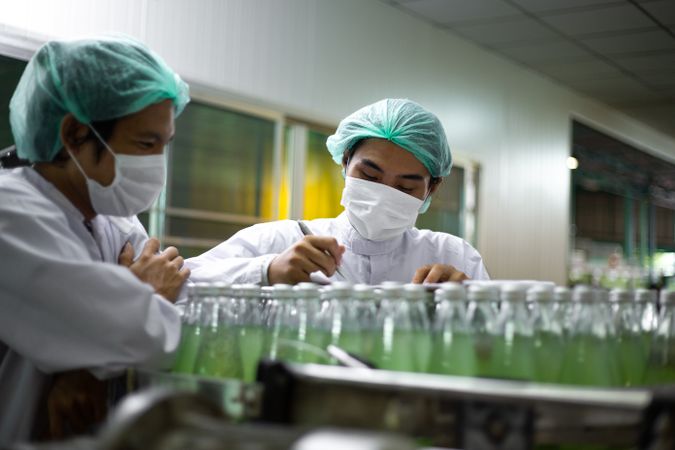 Male worker wearing protective uniform checking drink bottle on bottling machine following checklist in document.
