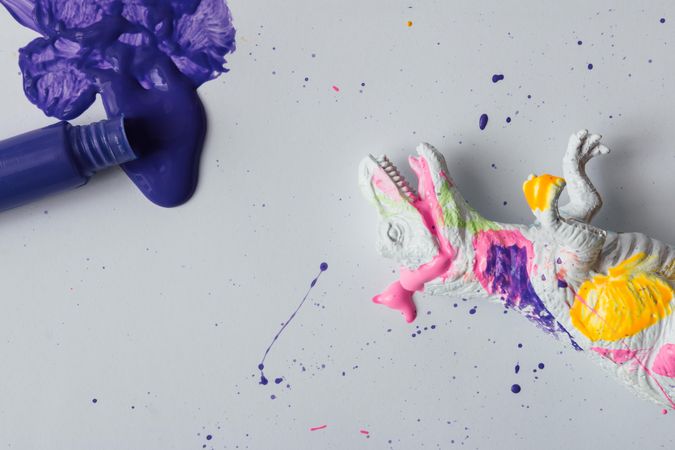 Plastic dinosaur splattered with different colors of paint lying down, with tube of paint