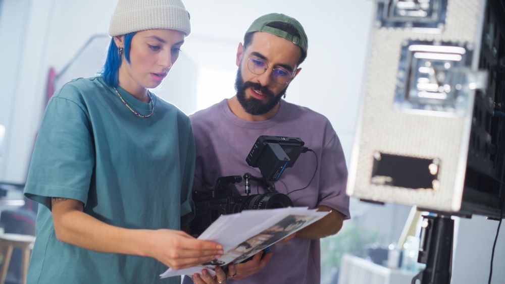 Young White Female And Middle Eastern Male Cinematographers Review A Script By A Camera On A Busy Film Set, Focusing On Video Production For A Documentary Or Tv Show.
