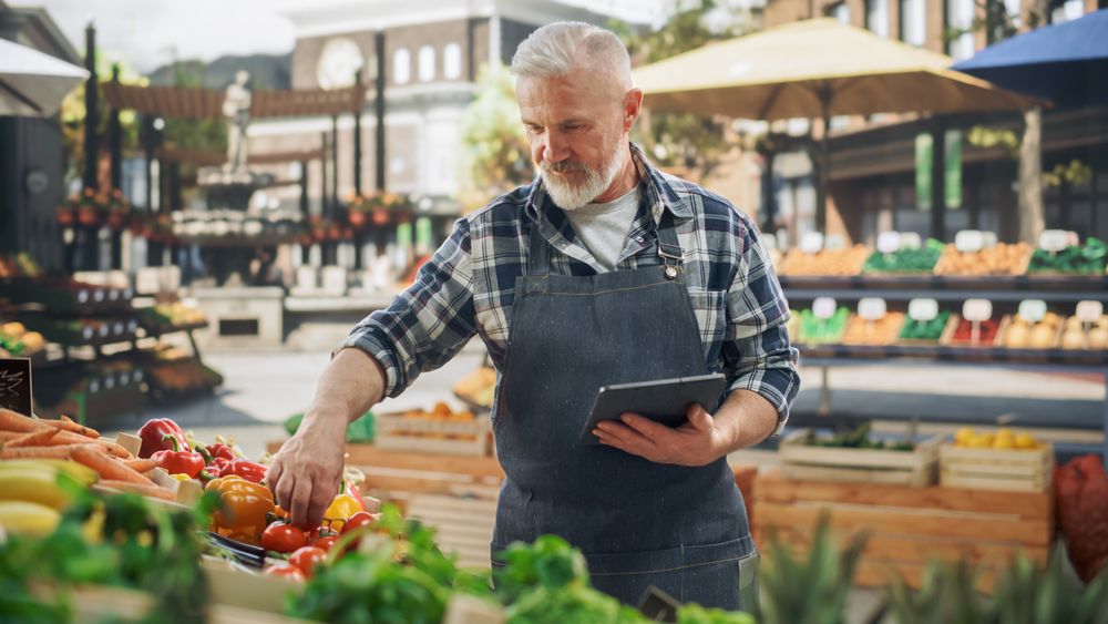 Handsome Adult Farmer Using Tablet Computer on a City Square with Vendors Selling Fresh Organic Agricultural