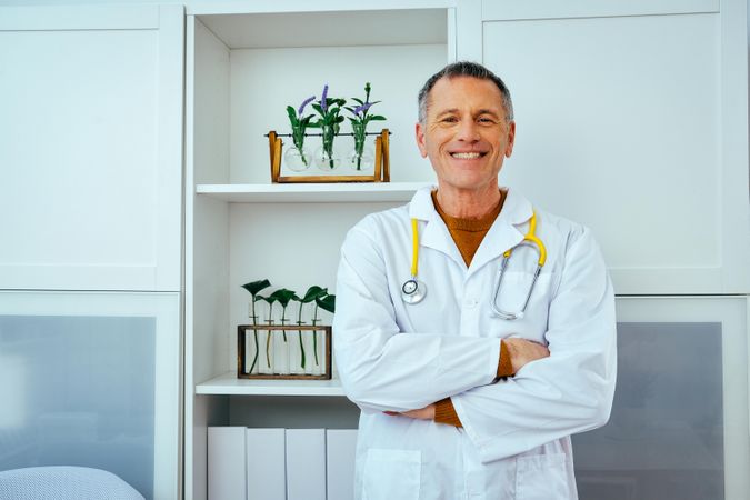 Older doctor standing in his office with arms crossed and smiling