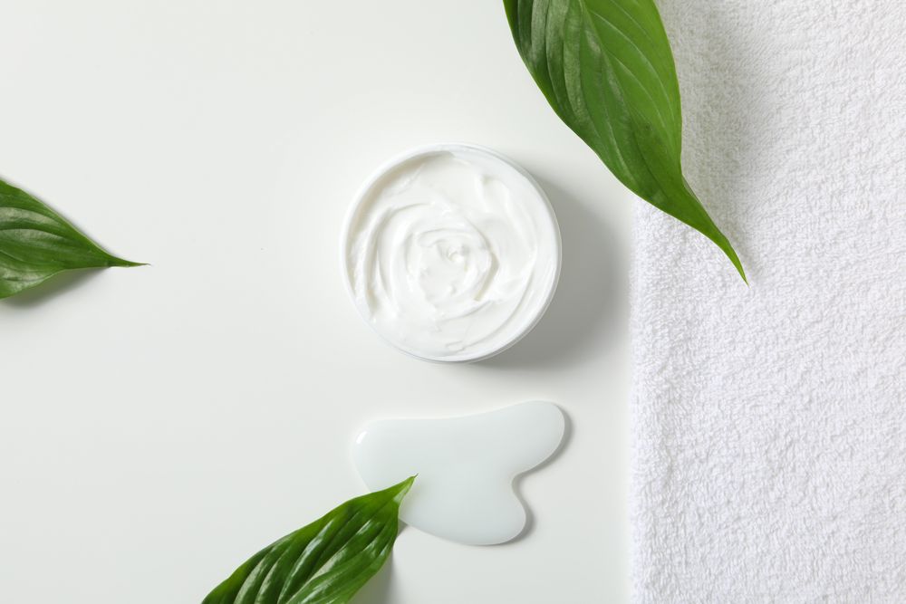 Jar with cream, towel and gua sha on light background, top view
