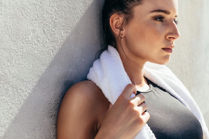Woman leaning to a wall with towel around her neck and looking away after a workout