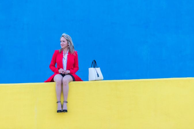 Woman in red coat sitting on bright yellow wall with blue in background with space for text