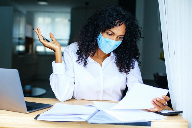 Woman frustrated while reading over documents with a laptop while wearing a facemask