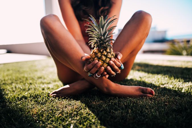 Tanned young woman sitting cross-legged holding a pineapple in the sun