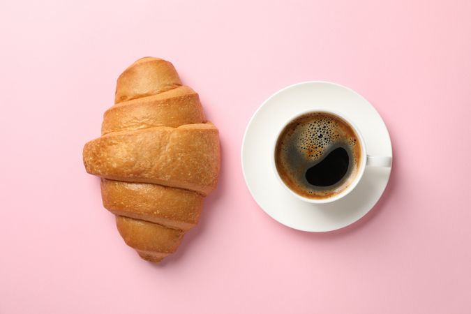 Croissant and cup of coffee on pink background, top view