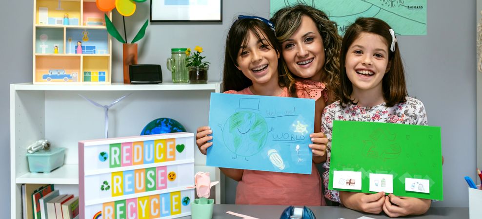 Teacher in classroom looking at camera next to students showing colorful ecological drawings