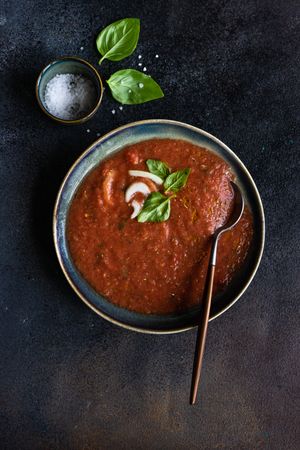 Top view of large bowl & spoon in Spanish gazpacho with basil leaf garnish and salt in blue bowl on counter