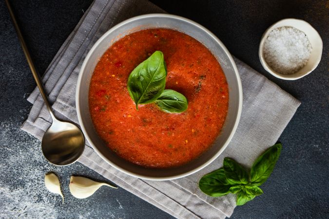 Top view of grey bowl of gazpacho soup with basil leaves on napkin