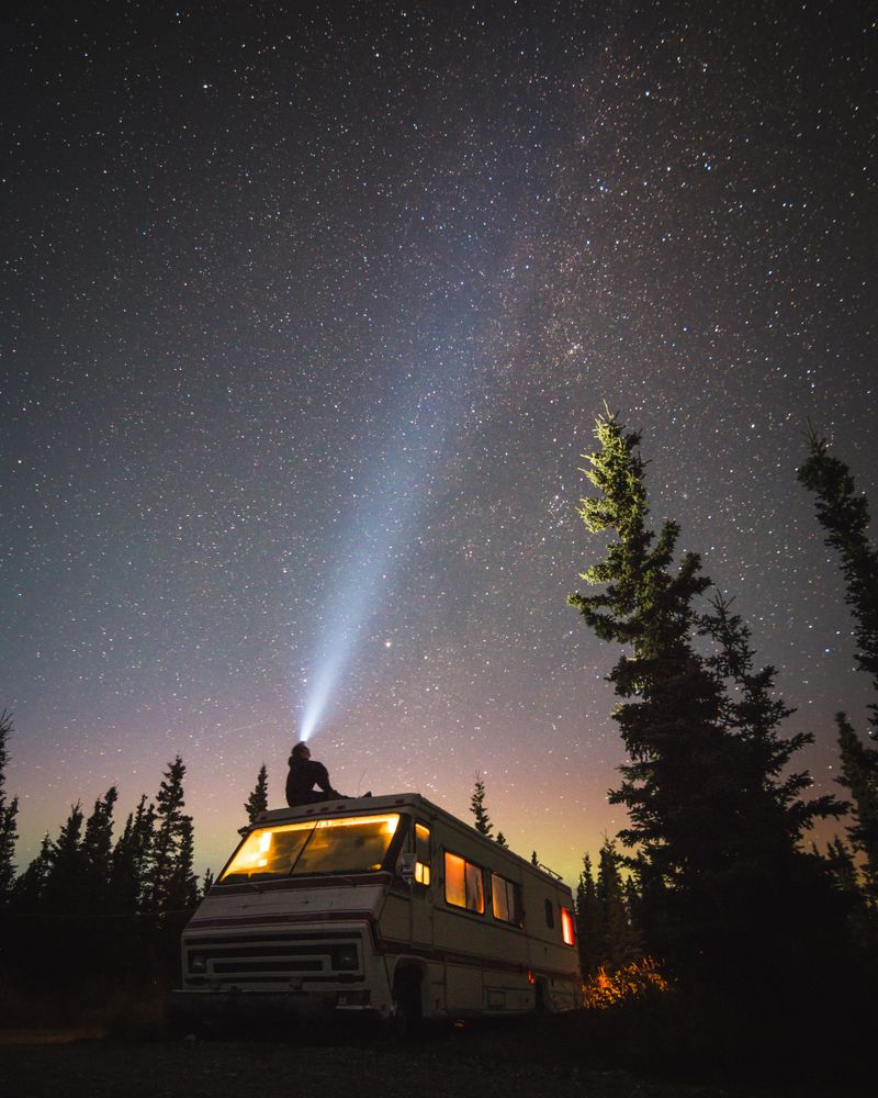 Silhouette of a person sitting on lit RV looking at stars in starry night sky