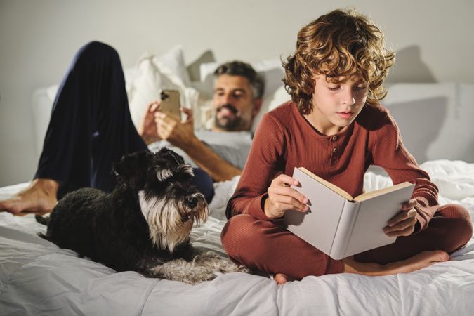 Boy sitting on bed and reading book against man browsing smartph
