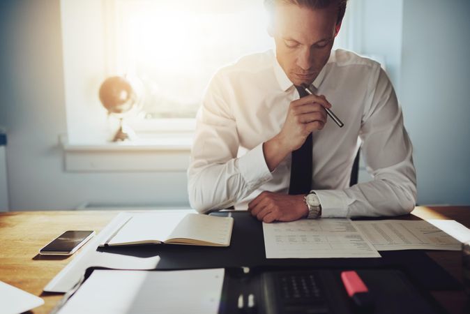 Man in shirt and tie sitting at desk reading over documents in a sunny office