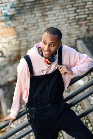 Happy male in overalls and pink hoodie leaning on railing of stairs outside