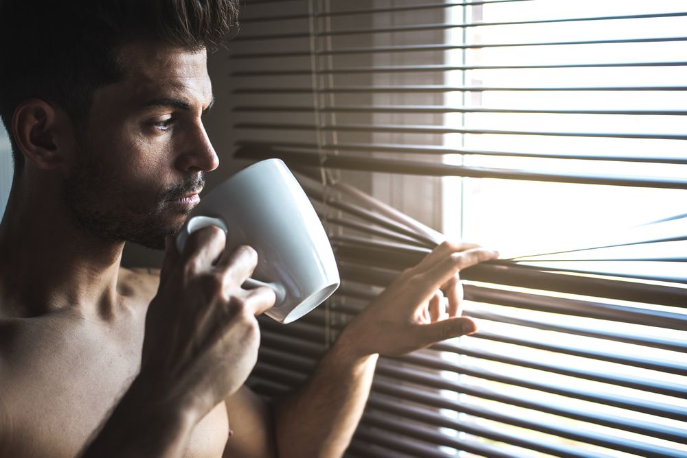 Man standing by a windows blind with a cup of coffee