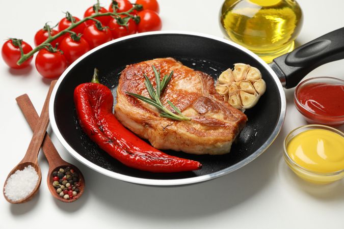 Pan with fried steaks and ingredients for cooking on blank background, close up