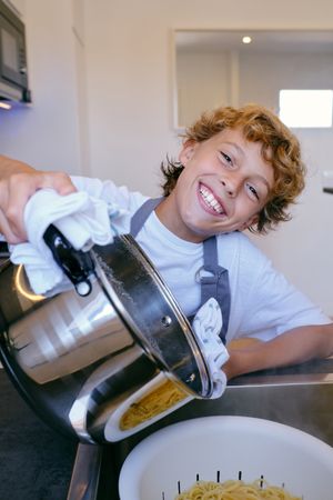 Cheerful boy with pot preparing spaghetti in kitchen