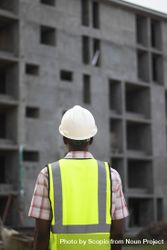 Back view of man wearing green vest and bump cap standing beside a building under construction 5oyRgb