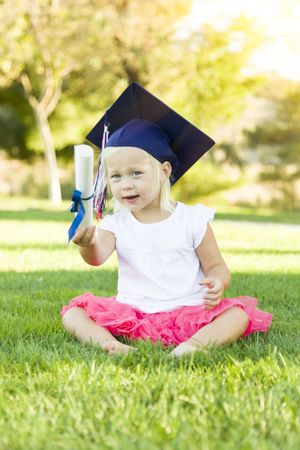 Little Girl In Grass Wearing Graduation Cap Holding Diploma With Ribbon