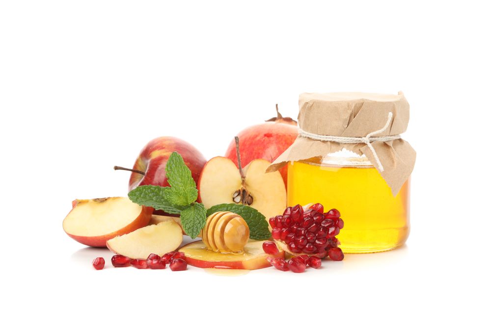 Honey dipper, pomegranate seeds, mint, and apple arranged on side of plain table